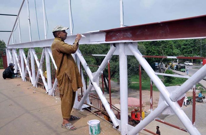 The painter paints the metal railings of the pedestrian bridge with white paint at Islamabad Expressway.