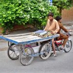 A person holding hand cart while sitting on rear seat of a motorcycle at General Bus Stand Road