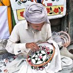 A skilled person making embroidery work on bed sheet and pillow to attract customer at his road side setup near NLI chowk