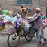 An elderly vendor along with bicycle loaded with household items for selling while shuttling in streets to earn livelihood.