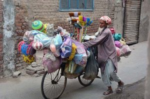 An elderly vendor along with bicycle loaded with household items for selling while shuttling in streets to earn livelihood. 