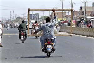 A person holding the frame of a traditional bed (charpai) while sitting on the rear seat of a motorcycle