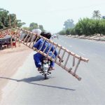 A person holding long ladder while sitting on the rear seat of a motorcycle on the way.