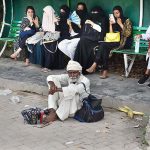 An elderly vendor waiting for customers to sell ballpoint pen while sitting in front of bus stop shed.