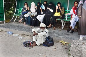 An elderly vendor waiting for customers to sell ballpoint pen while sitting in front of bus stop shed. 