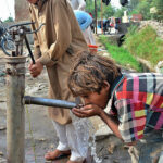 Children drinking water from hand pump outside road.
