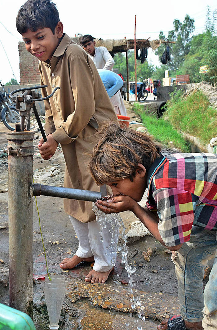 Children drinking water from hand pump outside road.