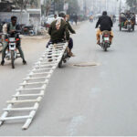 A person holding a ladder while sitting on the rear seat of a motorcycle near Dera Adda Chowk