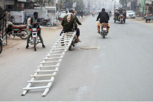 A person holding a ladder while sitting on the rear seat of a motorcycle near Dera Adda Chowk