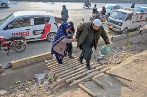 A person holding hand of woman to cross nullah through temporary wooden ladder along road at Pirwadhai.