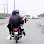 A pillion rider holding a pet goat on motorcycle traveling on a busy road.