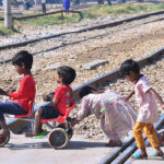 Children enjoy riding toy bicycle while crossing railway track near railway station.