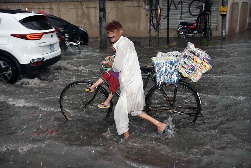 A person pushing his cycle sitting on a kid passing through rain water accumulated during heavy rain in the city.