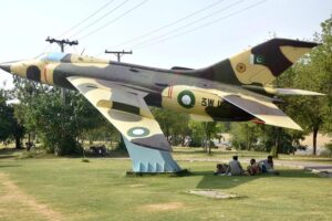 People sitting under the shadow of fighter plane model installed at roadside greenbelt in I-9 Sector during scorching hot weather in Federal Capital.
