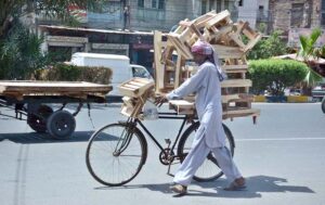 A street vendor pushes his bicycle, heavily laden with wooden stools, under the scorching sun on the city roads.