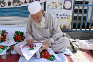 An aged person making embroidery work on the bed sheet and pillow to attract customer at his road side setup near Ramzan Hotel.