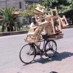 A street vendor pushes his bicycle, heavily loaded with wooden stools, under the scorching sun at the city road