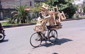 A street vendor pushes his bicycle, heavily loaded with wooden stools, under the scorching sun at the city road