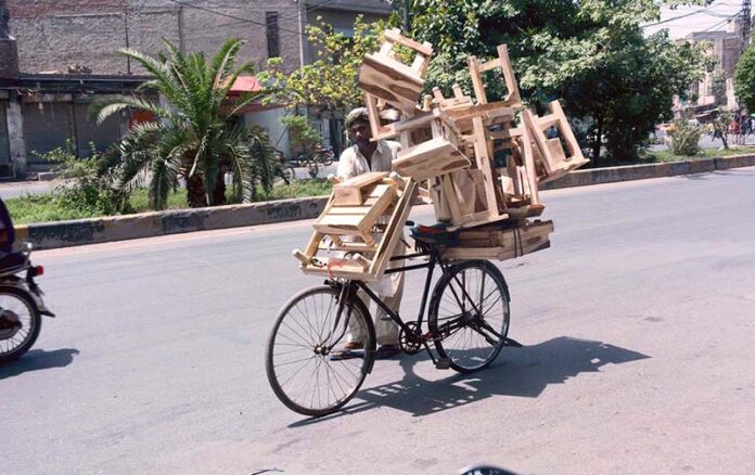 A street vendor pushes his bicycle, heavily loaded with wooden stools, under the scorching sun at the city road