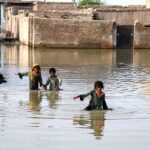 Children crossing flooded streets where rainwater has accumulated in front of houses at Qutb Colony