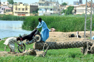 A man assists a motorcyclist in crossing sewerage water on a tree trunk bridge, highlighting the spirit of cooperation in asimabad.