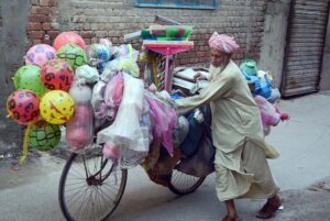 An elderly vendor rides his bicycle loaded with household items, shuttling through streets to earn his livelihood.
