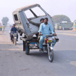 A tricycle rider transports the front body of a mini truck to a workshop, navigating with an unusual load in the city streets