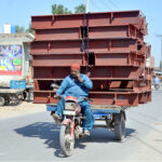 A tricycle holder on the way loaded with door frames while busy in the cell phone may cause any mishap