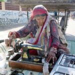 An elderly woman stitching clothes and selling kitchen items on footpath