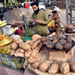 A hawker prepares roastred yams for sale while attracting customers with a colorful display of seasonal winter delights on his handcart near the Muslim Town Mor flyover