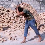 A laborer's back bowed under the weight of a heavy bundle of bricks as he carries it to the second floor during ongoing construction work
