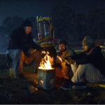 A group of people sitting around a crackling wood fire, seeking warmth and comfort as the winter chill envelops the Provincial Capital