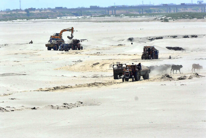A heavy machine operator loads mud onto a tractor trolley along the banks of the Indus River
