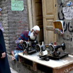 An aged man repairing the cloth sewing machine of a woman at his setup in a street of walled city