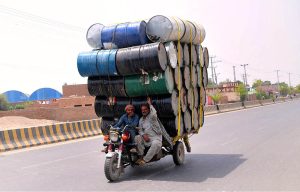 A motorcycle tri-cart loaded with dozens of empty oil drums, balanced carefully, makes its way through the city