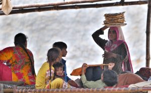 A nomadic woman carries chapatis on her head, walking towards her family waiting for lunch under a roadside hut