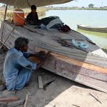 A carpenter busy repairs an old boat near a water pond.