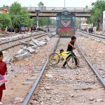 Children dangerously cross railway tracks in a residential area near railway tarck as a train approaches in the background, posing a serious risk of mishap urging immediate safety action by authorities
