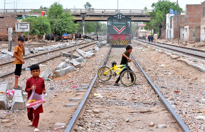 Children dangerously cross railway tracks in a residential area near railway tarck as a train approaches in the background, posing a serious risk of mishap urging immediate safety action by authorities