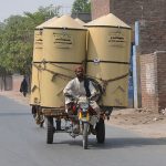 A tricycle holder dangerously overloaded with large wheat containers moves on the road, posing a serious risk of accident or mishap due to imbalance and poor visibility.