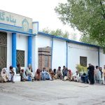 A large number of people sit outside a shelter waiting for food at the General Bus Stand