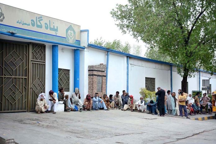 A large number of people sit outside a shelter waiting for food at the General Bus Stand