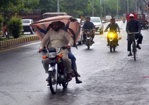 A motorcyclist shields himself with a cloth to stay dry while riding through light rain on a city road.