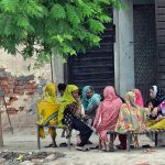 A group of women and children sit together on traditional woven cots (charpai) under the shade of a tree in a village setting, engaged in conversation. The scene captures a moment of community and togetherness, with vibrant, colorful clothing adding life to the rustic surroundings
