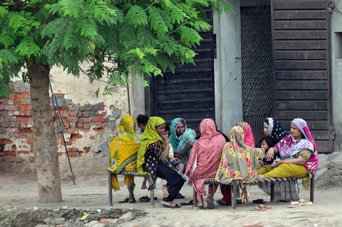 A group of women and children sit together on traditional woven cots (charpai) under the shade of a tree in a village setting, engaged in conversation. The scene captures a moment of community and togetherness, with vibrant, colorful clothing adding life to the rustic surroundings