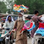 An elderly vendor carries colorful hand fans through a bustling market in search of customers, reflecting traditional street trade