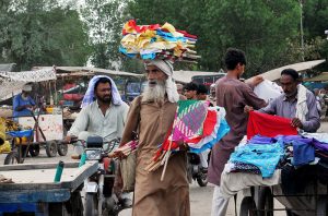 An elderly vendor carries colorful hand fans through a bustling market in search of customers, reflecting traditional street trade