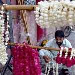 A vendor carefully arranges fresh flower garlands on a hanger, hoping to draw customers along a busy roadside near a city traffic junction