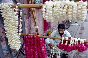 A vendor carefully arranges fresh flower garlands on a hanger, hoping to draw customers along a busy roadside near a city traffic junction