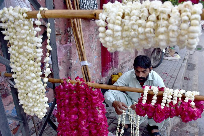 A vendor carefully arranges fresh flower garlands on a hanger, hoping to draw customers along a busy roadside near a city traffic junction